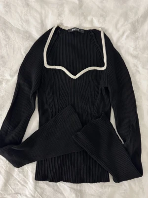 Svart ribbad topp Gina Tricot XS - Trendig svart ribbad topp från Gina Tricot med lång ärm och urringning kantad med vit detalj. Stretchigt material.