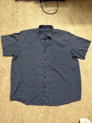 Blårutig kortärmad skjorta - Snygg kortärmad skjorta i mörkblått med vitt rutmönster. Skjortan har klassisk krage och knäppning framtill. Perfekt för dig som gillar en avslappnad men ändå stilren look.