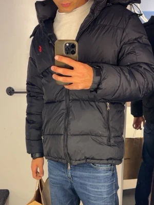 Svart pufferjacka från Polo Ralph Lauren - Svart pufferjacka från Polo Ralph Lauren med quiltad design och röd logga på bröstet. Jackan har dragkedja framtill, två sidofickor med dragkedja och hög krage. Perfekt för kalla dagar och ger en sportig look.