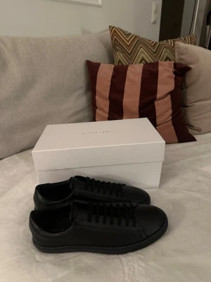 Svarta sneakers från Oliver Cabell - Stilrena helsvarta sneakers från Oliver Cabell i skinn med klassisk låg profil och rund tå. Skorna har svarta snören och platt sula, perfekta för en clean och modern look. Andvönda 1 gång 