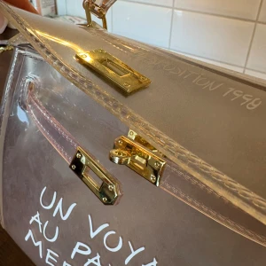 Transparent handväska med text - Säljer en transparent handväska med guldfärgade metalldetaljer. Väskan har vit text framtill med fransk text och stängs med ett klassiskt vridlås. Perfekt för dig som vill ha en unik och trendig accessoar.