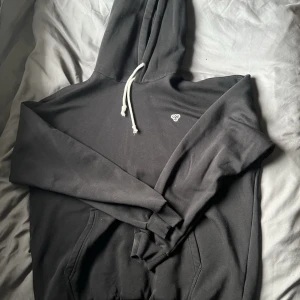 Svart hoodie med vit dragsko - Snygg svart hoodie med vit dragsko och liten broderad logga på bröstet. Klassisk känguruficka framtill och mjukt material som känns skönt mot huden. Perfekt för en avslappnad och stilren look.