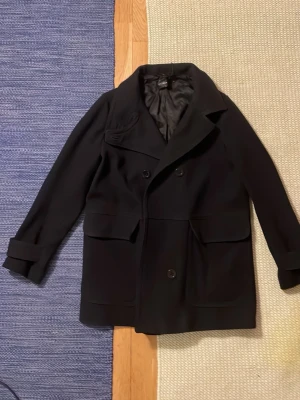 Svart skepparkavaj från Filippa K - Klassisk svart skepparkavaj från Filippa K med dubbelknäppning, bred krage och stora fickor framtill. Jackan är tillverkad i ett mjukt ullmaterial med slätt foder. Perfekt för dig som gillar stilrena och tidlösa plagg.  Storleken är L. Jackan är från början för kvinnor. Den passade mig och jag har vanligtvis storlek s eller m