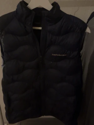Mörkblå dunväst Peak Performance - Säljer en mörkblå dunväst från Peak Performance, modell JR Helium Vest. Västen har hög krage, diskret logga på bröstet och dragkedja framtill. Perfekt för lager-på-lager och outdoor-vibes. Lätt och smidig att bära, passar till många olika stilar. Den har lite färgskvätt på sig o andra fläckar men kanske finns någon som vill ha en väst till ett billigt pris! Tveka inte på att ställa en fråga eller två😊😊