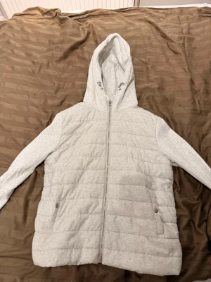 Ljusgrå quiltad hoodiejacka  - Säljer en ljusgrå quiltad hoodiejacka med dragkedja framtill och två fickor med knappstängning. Jackan har långa ärmar och en stor huva. Perfekt för dig som gillar en enkel och clean stil.