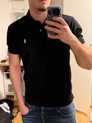 Svart pikétröja från Polo Ralph Lauren - Klassisk svart pikétröja från Polo Ralph Lauren med vit broderad logga på bröstet. Tröjan har krage, knappar framtill och korta ärmar. Tillverkad i mjuk bomull som känns skön mot huden.