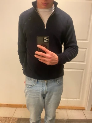 Mörkblå stickad tröja med halvzip - Snygg mörkblå stickad tröja med halvzip och hög krage. Tröjan har långa ärmar och ribbade muddar vid ärmslut och nederkant. Perfekt för lager på lager och passar till både jeans och chinos.