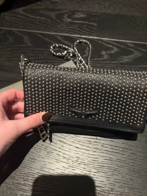 Svart kuvertväska Zadig & Voltaire - Snygg svart kuvertväska från Zadig & Voltaire med silverfärgade nitar över hela framsidan och en kedjerem. Väskan är gjord i skinn och har en liten logga framtill samt dragkedja. Perfekt för dig som vill ha en edgy accessoar.