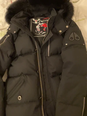 Svart pufferjacka Moose Knuckles M - Säljer en svart pufferjacka från Moose Knuckles med stor fluffig pälskant på huvan och snygga metalldetaljer. Jackan har dragkedjor, flera fickor och broderad insida med hockeymotiv. Perfekt för kalla dagar och riktigt stilren. Den är äkta och finns inga defekter 