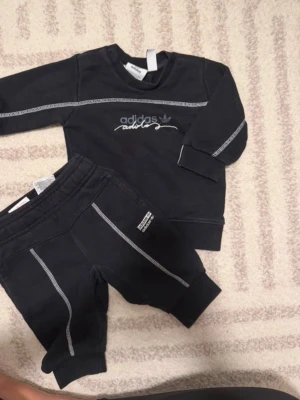 Svart Adidas babyset 3-6 mån - Säljer ett svart Adidas set för baby, storlek 3-6 månader. Tröjan har vita kontrastsömmar, Adidas-logga och tryck framtill samt muddar med ränder. Byxorna har matchande sömmar och liten Adidas-patch på benet. Perfekt för en sportig look!Adidas baby set size 3-6m Slightly washed out but in perfect condition otherwise   Smoke-free & pet-free home.