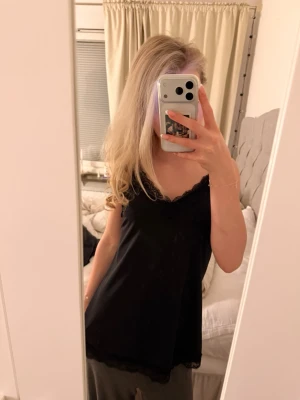 Svart spetsdetaljerad linne - Säljer en svart linne med tunna axelband och v-ringad halsringning. Linnet har fina spetsdetaljer längs urringningen och nederkanten som ger en extra touch. Modellen är loose fit och materialet känns mjukt och följsamt.