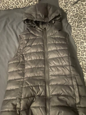 Svart dunväst med huva från ONLY - Snygg svart dunväst från ONLY med quiltad design och dragkedja framtill. Västen har en stor huva och är perfekt för lager på lager-stil. Tillverkad i ett glansigt material som ger en modern look. Passar perfekt till höst och vinter för en trendig outfit.