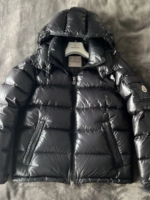 Svart dunjacka från Moncler - Snygg svart dunjacka från Moncler med quiltad design och huva. Jackan har två dragkedjeförsedda fickor framtill och en Moncler-logga på ärmen. Perfekt för kalla dagar och ger en stilren look.