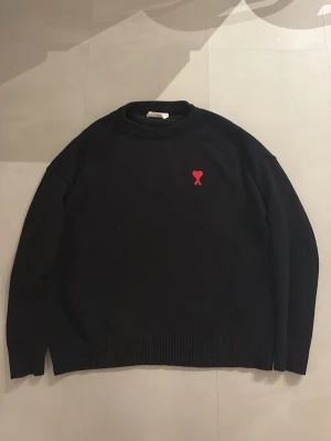 Svart stickad tröja från Ami Paris - Klassisk svart stickad tröja Ami:s ikoniska röda hjärtemblem. Men har en syning på armen som ni kan se på sista bilden 