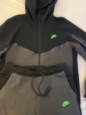 Nike Tech tracksuit - Nike Tech Fleece hoodie och mjukisbyxor i svart och grått med dragkedja och huva. Tröjan har en neongrön Nike-logga på bröstet och är tillverkad i ett mjukt och sportigt material. Perfekt för en streetwear-look eller till träningen.                          Nypris: 2500kr. Kvitto finns