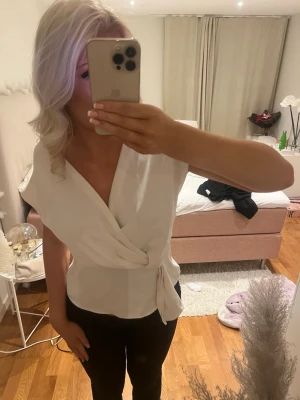 Vit omlottopp från Zara - Säljer en vit omlottopp från Zara med korta ärmar och knyt framtill. Toppen har en djup v-ringning och är gjord i ett mjukt, lätt material som ger ett snyggt fall. Perfekt att styla med jeans eller kjol för en fräsch look.