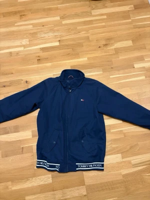Mörkblå overshirt från Tommy Hilfiger - Snygg mörkblå overshirt från Tommy Hilfiger med dragkedja framtill, två fickor med knapp och logga på bröstet. Jackan har ribbade muddar med Tommy Hilfiger-tryck och meshfoder på insidan för extra komfort. Perfekt för blåsiga dagar. Passar S, aldrig använd, inga skador eller sådant!