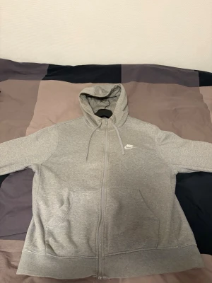 Grå zip hoodie från Nike - Snygg grå hoodie från Nike med dragkedja framtill och klassisk vit Nike-logga på bröstet. Hoodien har huva med dragsko och två fickor framtill. Tillverkad i mjukt sweatshirtmaterial som är perfekt för chill eller träning. Storlek M, inga skador. Jättefint skick.