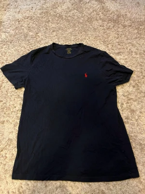 Svart t-shirt från Polo Ralph Lauren - Klassisk svart t-shirt från Polo Ralph Lauren med röd broderad logga på bröstet. T-shirten har rund halsringning och korta ärmar. Tillverkad i mjuk bomull och har en normal passform som sitter snyggt på kroppen.