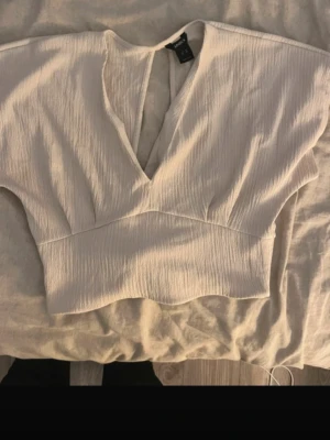 Ljusvit croppad omlottopp Lindex  - Säljer en ljusvit croppad omlottopp från lindex med djup v-ringning och knyt i ryggen. Toppen har korta ärmar och är gjord i ett mjukt, lätt strukturerat tyg som känns luftigt. Perfekt för dig som gillar trendiga och stilrena plagg.