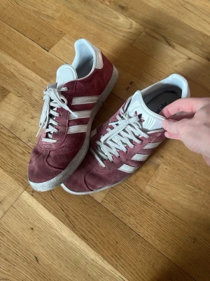 Adidas Gazelle vinröda sneakers - Säljer ett par vinröda Adidas Gazelle sneakers i mocka med vita detaljer och klassiska tre ränder på sidan. Skorna har snörning, vit platt sula och Adidas-logga på hälen. Perfekta för dig som gillar retrostil och snygga färgkombinationer.