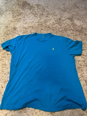 Blå t-shirt från Polo Ralph Lauren - Klassisk blå t-shirt från Polo Ralph Lauren med korta ärmar och rund hals. T-shirten har den ikoniska lilla broderade loggan i gult på bröstet. Tillverkad i mjuk bomull för en skön känsla och avslappnad passform.