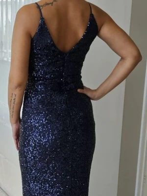 Mörkblå paljettklänning omlott - Snygg mörkblå klänning med paljetter och omlott-design. Klänningen har smala axelband, v-ringning och en slits framtill. Perfekt för dig som vill sticka ut med glitter och elegant look.
