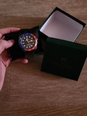  klocka med blå och röd bezel - Snygg  klocka med svart urtavla, blå och röd bezel samt silverfärgad länk i metall. Klockan har tydliga markeringar och klassisk design. Levereras i original grön Perfekt accessoar för dig som gillar exklusiv stil.