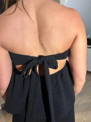Svart bandeau topp med knyt i ryggen - Säljer en svart bandeau topp med volang och snygg knytning i ryggen. Toppen är axelbandslös och har en luftig, lite lös passform. Perfekt för dig som vill ha en enkel men ändå trendig look.