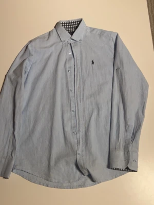 Ljusblå linneskjorta Polo Ralph Lauren - Säljer en ljusblå slim fit skjorta från Polo Ralph Lauren i 100% linne. Skjortan har klassisk krage, knappar framtill och lång ärm. Insidan av kragen och manschetterna har snyggt rutigt mönster i svart och vitt. Perfekt för dig som gillar stilrena detaljer.