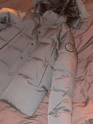 Svart dunjacka med huva från canada goose - Snygg svart dunjacka från canada goose med huva och märke på ärmen. Jackan har quiltad design, tryckknappar och dragkedja framtill. Perfekt för kalla dagar och har en klassisk pufferstil.