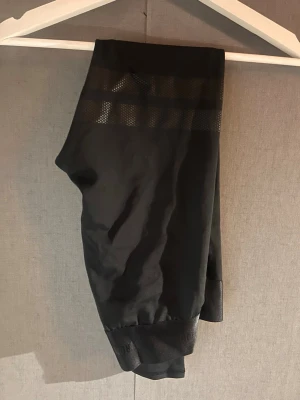 Svarta leggings med meshdetalj - Säljer ett par svarta leggings/träningtajts med snygg meshdetalj upptill. De har en tight passform och är perfekta för träning eller chill. Materialet känns stretchigt och bekvämt, och designen är enkel men modern.