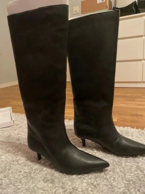 Svarta höga stövlar i skinn - Säljer ett par svarta höga stövlar med spetsig tå och låg klack. De är tillverkade i slätt skinn och har en elegant, tidlös look som passar till många olika outfits. Perfekta för dig som vill ha en clean och stilren vibe. Endast testade inomhus, passade tyvärr inte.