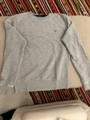 Grå sweatshirt från GANT, storlek S - Klassisk grå sweatshirt från GANT i storlek S. Tröjan har rund halsringning, ribbade muddar vid ärmslut och nederkant samt en liten broderad GANT-logga på bröstet. Perfekt basic-plagg för lager på lager och chill stil. Lite urtvättning vid handlederna. 