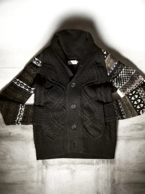 Svart kofta med mönstrade ärmar - Unik svart kofta med grovstickat mönster framtill och knappar. Ärmarna har flera olika mönster i svart, vitt och grått som ger en cool kontrast. Perfekt för dig som vill sticka ut lite extra under höst och vinter.