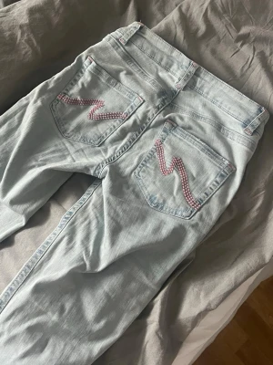 Ljusblå ripped jeans från Nelly - Säljer ett par ljusblå jeans från Nelly med slitningar på knäna och snygga rosa detaljer på bakfickorna. Klassisk femficksmodell med normal passform och utsvängda ben. Här sprättat upp sömmen där nere och jeansen är slittna där bak då jag gått på dem.