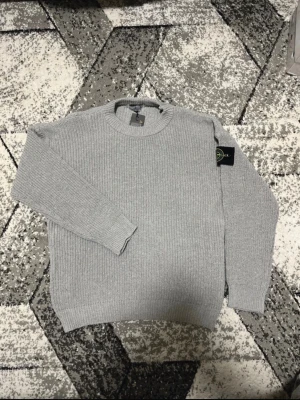 Grå stickad tröja Stone Island - Säljer en grå stickad tröja från Stone Island i storlek M. Tröjan har rund hals, ribbade muddar och den klassiska Stone Island-loggan på ärmen. Perfekt för dig som vill ha en clean och stilren look med premiumkänsla.
