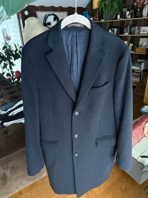 Mörkblå kashmir och ull rock från Oscar Jacobson - Säljer en klassisk mörkblå rock från Oscar Jacobson, modell Savile Row Coat. Rocken har tre knappar framtill, diskreta fickor och elegant krage. Insidan är fodrad med mörkblått och brunt tyg, vilket ger en lyxig känsla. Perfekt för dig som vill ha en stilren och tidlös look. Rocken är gjort utav 10% kashmir 60% ull 30% polyamide. Rocken är i ny skick samt kostar 6000kr ifrån butik 