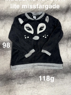 Svart tröja med djuransikte och grå detaljer - Mysig svart tröja med ett gulligt djuransikte framtill och grå ribbade muddar vid ärmslut, hals och nederkant. Perfekt för höst och vinter, och tillverkad i mjukt material som känns skönt mot huden. Långärmad modell som passar till vardags.🌸 Muddarna har blivit lite missfärgade. Storlek 98 
