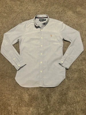 Ljusblå skjorta från Polo Ralph Lauren - Säljer en stilren ljusblå skjorta från Polo Ralph Lauren i slim fit. Skjortan har klassisk krage, långa ärmar och knappar hela vägen. Broderad logga på bröstet och tillverkad i mjuk bomull. Perfekt för dig som gillar en clean och tidlös look.