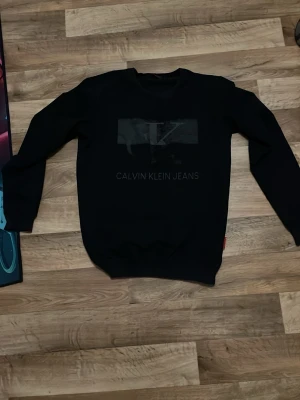 Svart sweatshirt Calvin Klein Jeans L - Säljer en svart sweatshirt från Calvin Klein Jeans i storlek L. Tröjan har ett diskret tryck med CK-logga och text på bröstet. Klassisk rund halsringning och långa ärmar. Perfekt för dig som gillar stilrena och enkla plagg.