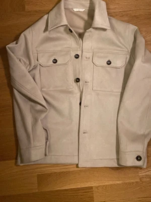 Beige overshirt från Zara i mockaimitation - Säljer en beige overshirt från Zara i mockaimitation. Jackan har två bröstfickor med knappar, klassisk krage och knäppning framtill. Perfekt lager-på-lager-plagg med stilren och enkel design.