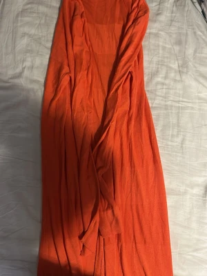 Lång orange röd kofta från Sincerely Loved - Lång, öppen kofta i en stark orange röd färg från Sincerely Loved. Koftan är tunnstickad, har långa ärmar och ett skönt flowy fall. Perfekt att slänga över en topp för extra färg och stil. Passar dig som gillar statement-plagg och vill ha något bekvämt men ändå snyggt.