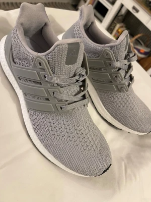 Grå Adidas Ultraboost sneakers - Säljer ett par grå Adidas Ultraboost sneakers med stickad ovandel och vita Boost-sulor. Skorna har snörning, plastdetaljer på sidorna och svart yttersula från Continental. Perfekta för dig som vill ha både stil och komfort.