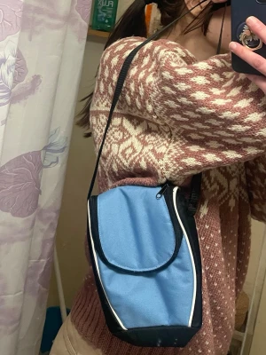 Blå axelväska med vit kant - Säljer en blå kameraväska med svart justerbar rem och vita detaljer längs kanterna. Väskan har dragkedja upptill och ett rymligt fack med vit insida. Perfekt storlek för kamera eller bara för vanliga accessoarer och enkel att bära crossbody.