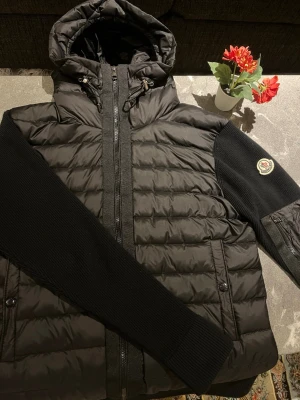 Moncler Cardigan - Säljer en svart Moncler dunjacka med quiltad kropp och stickade ärmar. Jackan har huva med dragsko, dragkedja framtill och fickor med knappstängning. Klassisk Moncler-logga på ärmen och snygga detaljer som ger en modern look.