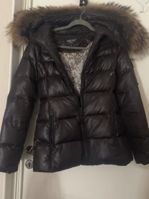 Brun pufferjacka med fuskpäls från Hollies - Säljer en brun pufferjacka från Hollies med snygg fuskpäls på huvan och blommigt mönstrat innerfoder. Jackan har dragkedja framtill, två fickor med dragkedja och en skön, varm känsla. Perfekt för kalla dagar och riktigt stilren look.