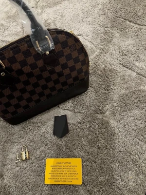 Antonio Vuitton rutig handväska brun - Säljer en ikonisk handväska från  Vuitton i klassiskt brunt och mörkbrunt rutigt mönster. Väskan har guldfärgade metalldetaljer, dragkedja och medföljande lås och nyckel. Tillverkad i slitstarkt skinn och har en snygg bågformad design.