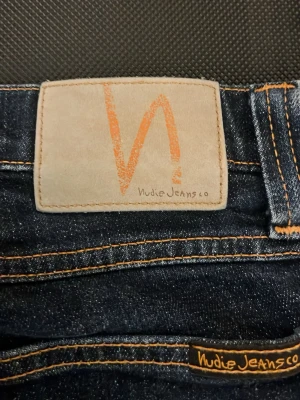 Nudie Jeans blå jeansbyxor W28 L32 - Säljer ett par klassiska blå jeans från Nudie Jeans med snygga kontrastsömmar och ikonisk broderad bakficka. Modellen har normal passform och raka ben. Tillverkade i slitstarkt bomullsmaterial, perfekta för en avslappnad och stilren look.