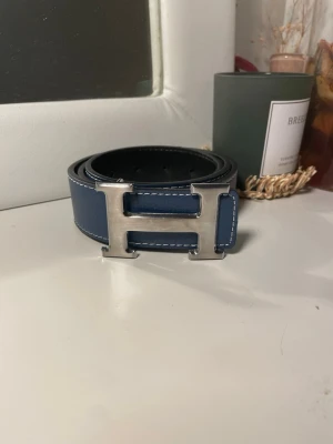 Blått bälte med H-spänne - (priset kan diskuteras) Snyggt blått bälte i skinn med ett stort, silverfärgat H-spänne. Bältet har vit kontrastsöm längs kanterna och flera hål för justerbar passform. Perfekt accessoar för att lyfta vilken outfit som helst.